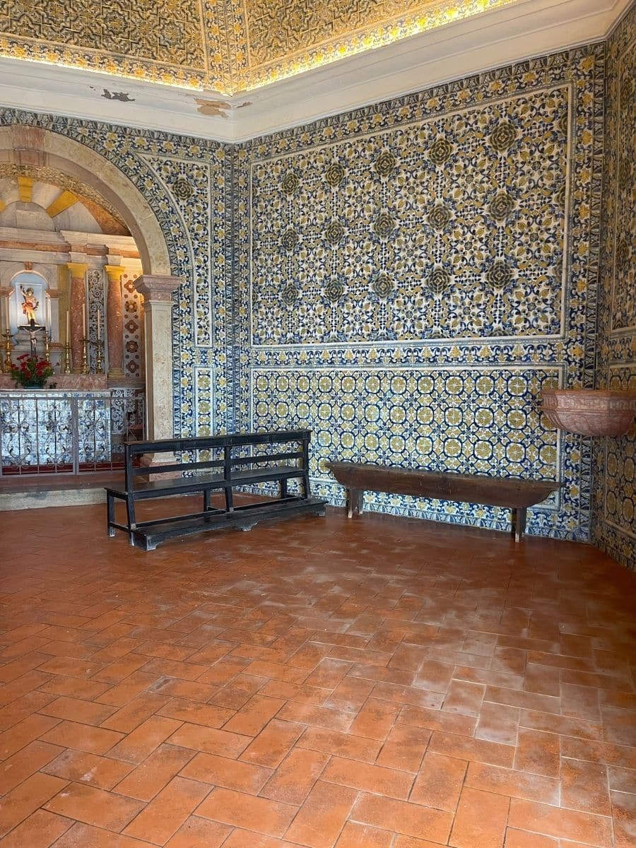 Interior of a small chapel richly decorated with traditional Portuguese azulejo tiles in blue, yellow, and white floral patterns covering the walls and part of the ceiling. A simple wooden bench and a black metal prayer kneeler sit on a terracotta-tiled floor. In the background, an ornate altar with a statue of a saint and flower arrangements is framed by a stone arch.