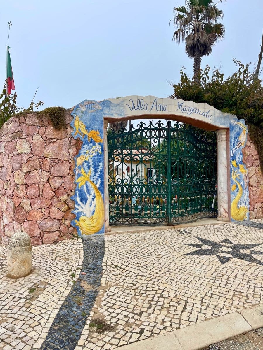 The entrance to Villa Ana Margarida in Ericeira, Portugal, featuring a decorative wrought iron gate framed by a colorful tiled archway. The arch is adorned with blue and yellow azulejo tiles depicting sea creatures and mermaids. The surrounding wall is made of rustic pink stone, complementing the traditional Portuguese cobblestone pavement in the foreground. A palm tree towers above the entrance, adding to the coastal charm. The Portuguese flag waves gently in the background, hinting at the villa's local heritage.