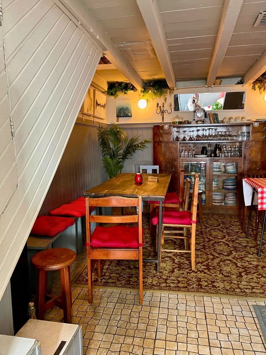 A cozy, rustic dining area inside a traditional European restaurant. The space features a wooden table with red-cushioned chairs, a vintage wooden cabinet filled with glassware and plates, and a warm, dimly lit ambiance. The slanted ceiling and wooden beams add charm, while greenery and small decorative elements enhance the inviting atmosphere. The combination of tiled and carpeted flooring further emphasizes the homey, old-world aesthetic.
