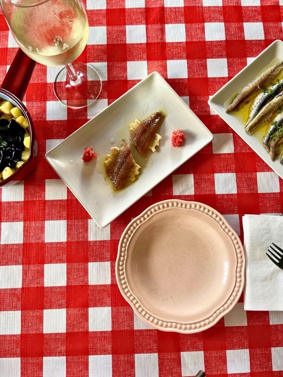 A red and white checkered tablecloth with a selection of tapas dishes and a glass of white wine. In the center, a white rectangular plate holds two pieces of anchovy fillets on crackers, garnished with a dollop of red sauce. To the right, another plate features marinated anchovies in olive oil with herbs. In the top left corner, a small pan contains fried potatoes topped with a dark sauce. A pink ceramic plate with a decorative rim sits empty, alongside a neatly folded napkin with a fork. The setting suggests a rustic, cozy dining experience.