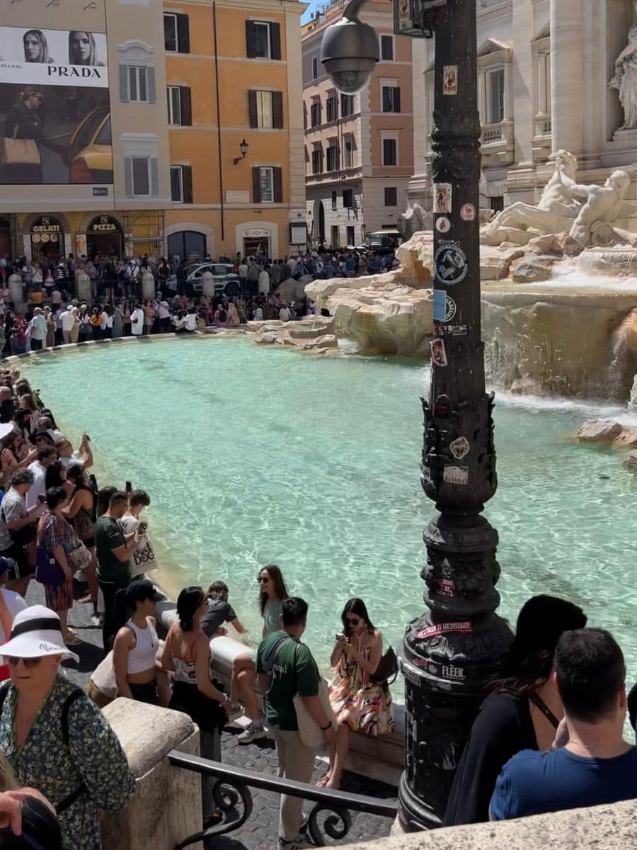 Crowded Trevi Fountain in Rome with tourists, a sticker-covered lamppost, and colorful buildings in the background.