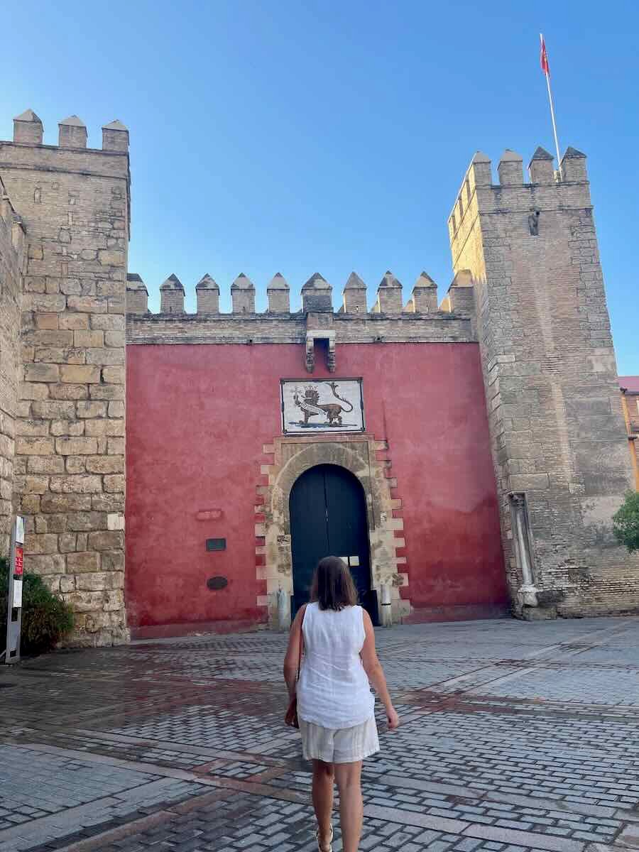 A person walking toward the main entrance of the Alcázar of Seville. The entrance is flanked by large, crenelated stone walls and a red-painted section with a black arched door, above which is a coat of arms featuring a lion.