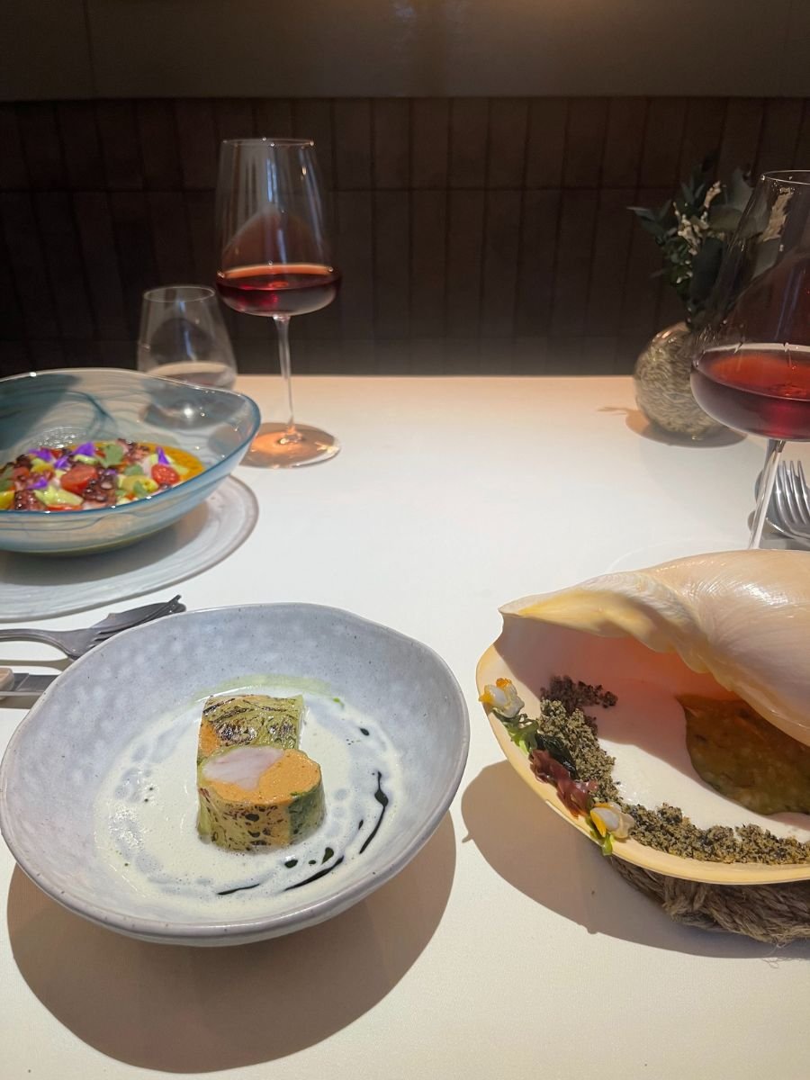 A fine-dining table view showing two artfully plated starters—a colorful octopus salad in a glass bowl and a creamy soup with a green-herb roulade—accompanied by two stemmed glasses of rosé and a small floral arrangement in the background.