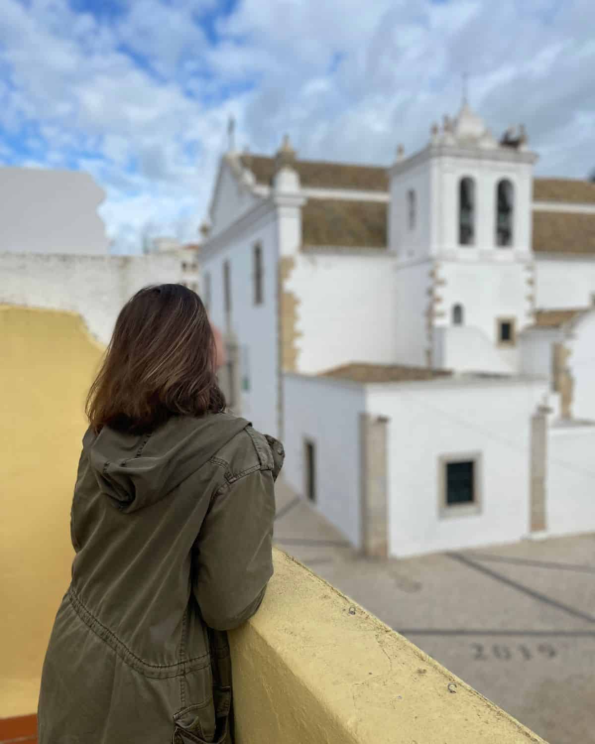 A woman with shoulder-length brown hair, wearing an olive-green jacket, is standing on a balcony overlooking a white building with a traditional architectural style and a bell tower under a partly cloudy sky.