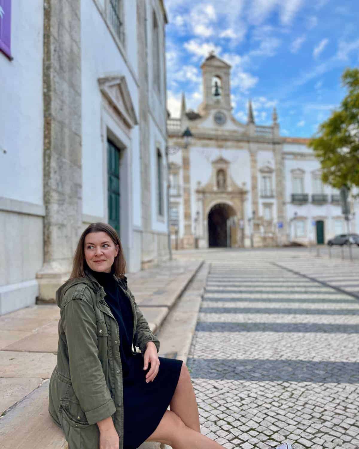The female traveler sits casually on a stone ledge in a plaza, smiling slightly, with the same historic building featuring a bell tower in the soft-focused background, under a partly cloudy sky.