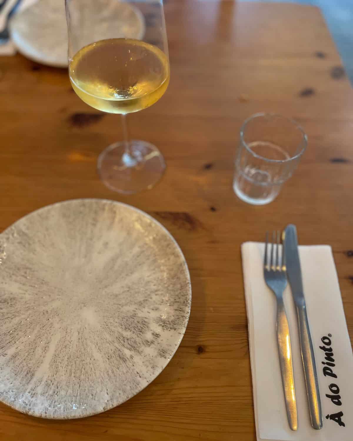 A rustic wooden dining table is set for one, with a white plate, cutlery wrapped in a napkin, and a glass of white wine that captures the relaxed ambiance of the eatery. The table's natural wood grain adds a warm, inviting tone to the scene.