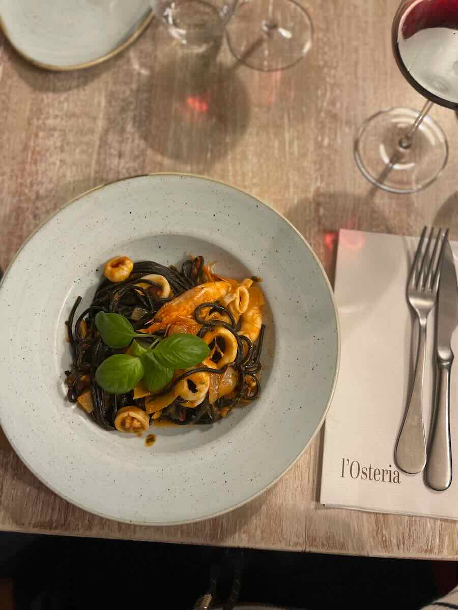 A plate of squid ink pasta with seafood, garnished with fresh basil, served at l'Osteria in Faro. The table is set with wine glasses and cutlery, creating an elegant dining ambiance.