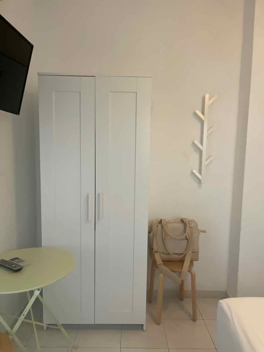 A simple interior of a room in Faro, featuring a white wardrobe, a light green round table with a TV remote, and a wooden chair holding a beige bag. A modern coat rack is mounted on the wall.
