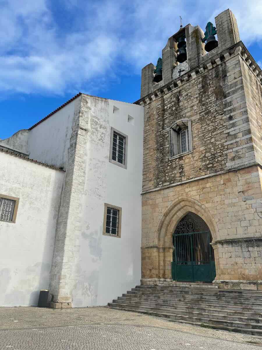 The bell tower of Faro Cathedral in Portugal, featuring a Gothic arched entrance, stone walls, and visible bells, with a white adjoining building and a vibrant blue sky in the background.