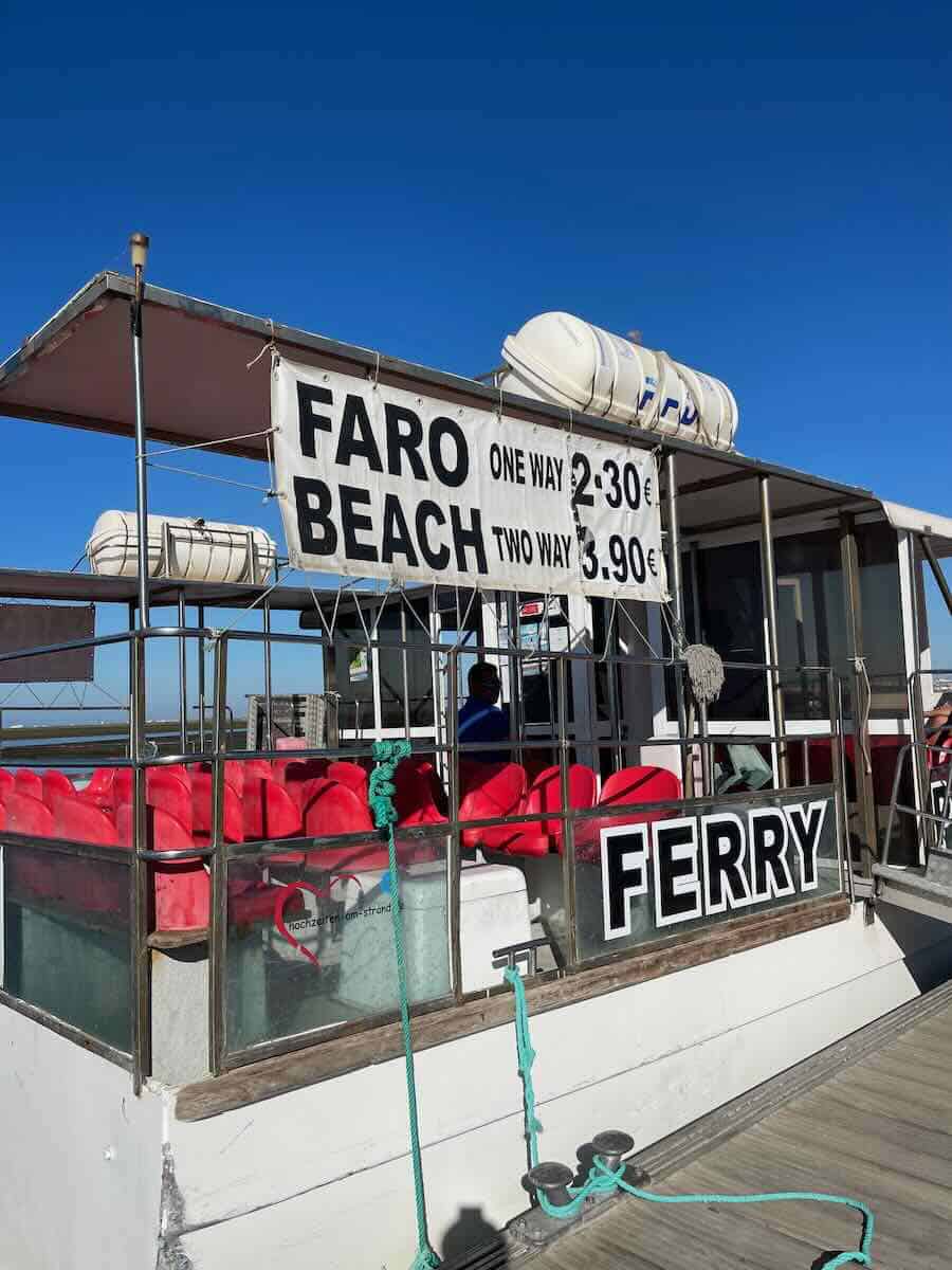Ferry at Faro Beach with a sign displaying ticket prices: one-way for €2.30 and round-trip for €3.90, against a clear blue sky.