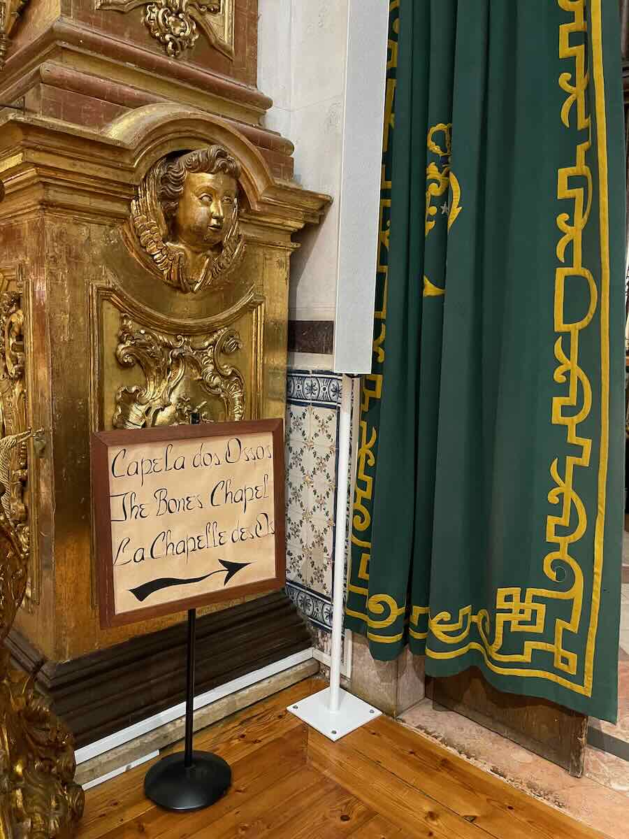 A detailed interior view in Faro, showcasing a gilded decorative carving of an angel and a wooden sign pointing toward the "Capela dos Ossos" (The Bones Chapel), framed by green and gold curtains with ornate trim.