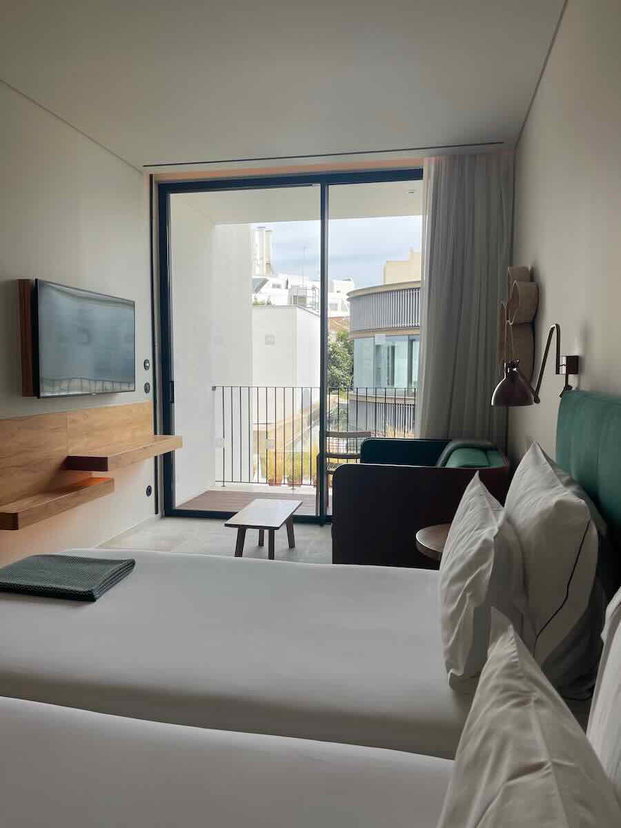 A stylish hotel room in Corfu with twin beds, a wall-mounted TV, a small table and seating area, and a balcony overlooking modern architecture.