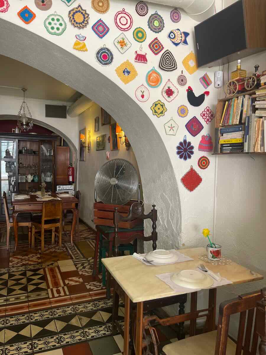 A cozy restaurant interior with a colorful wall of crocheted ornaments, wooden tables and chairs, and a tiled floor.