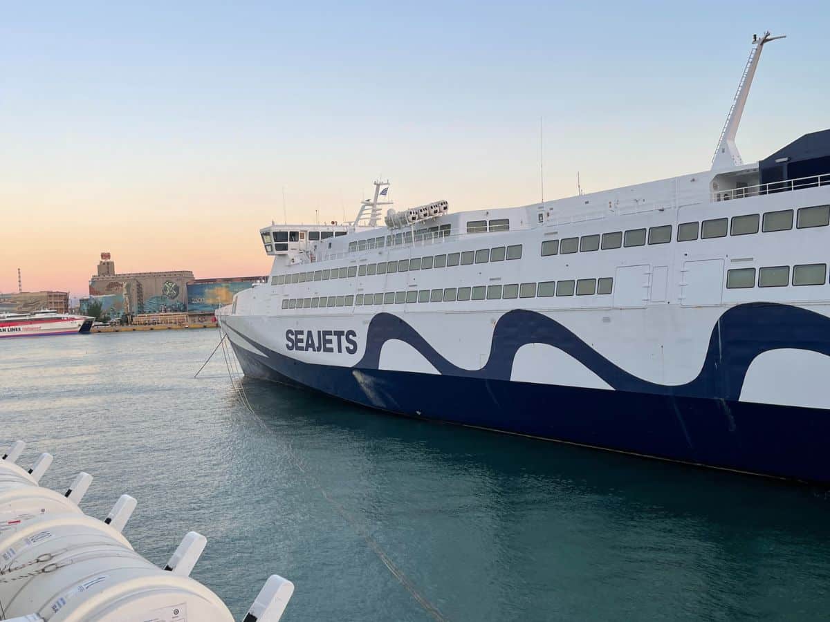 Seajets ferry docked at a port, with a white and blue wave design along its side. The scene is set against a soft sunset sky with industrial buildings and another ferry in the distance across the water.