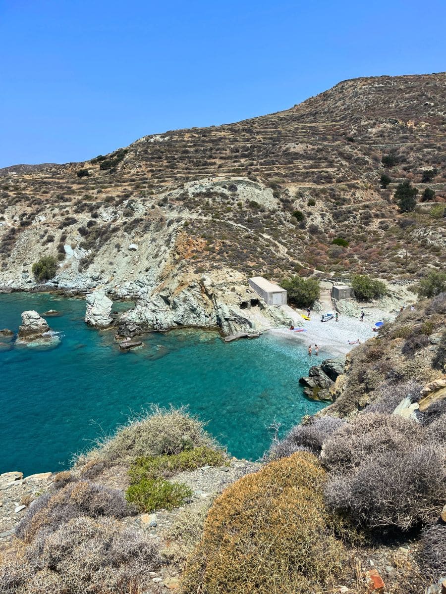 A secluded cove on Folegandros island, Greece, with clear turquoise waters gently meeting a small pebble beach. A few sunbathers and swimmers enjoy the peaceful surroundings. Jagged rock formations emerge from the sea, adding to the dramatic coastal scenery. Above the cove, terraced hills with sparse vegetation stretch under a cloudless blue sky, creating a striking contrast between the arid landscape and the inviting waters.