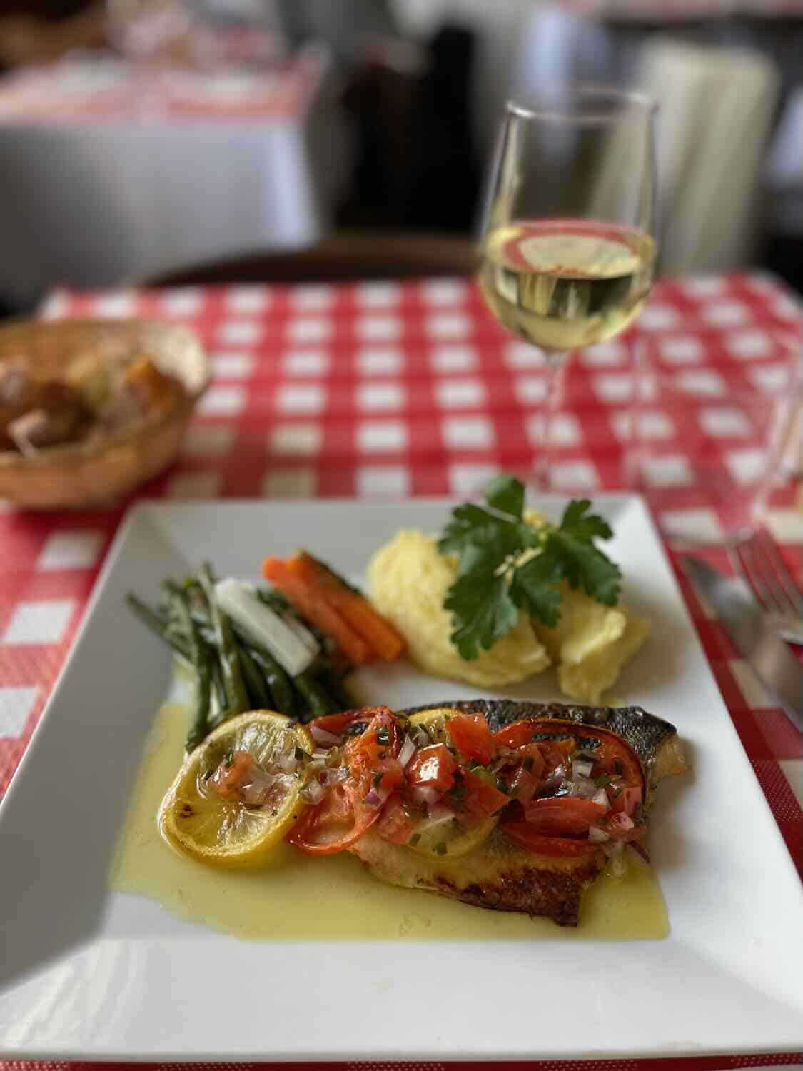 A gourmet meal featuring trout topped with a tomato and lemon sauce, served alongside a glass of white wine, green beans, carrots, and mashed potatoes on a table with a red and white checkered tablecloth, embodying a classic Parisian dining experience.