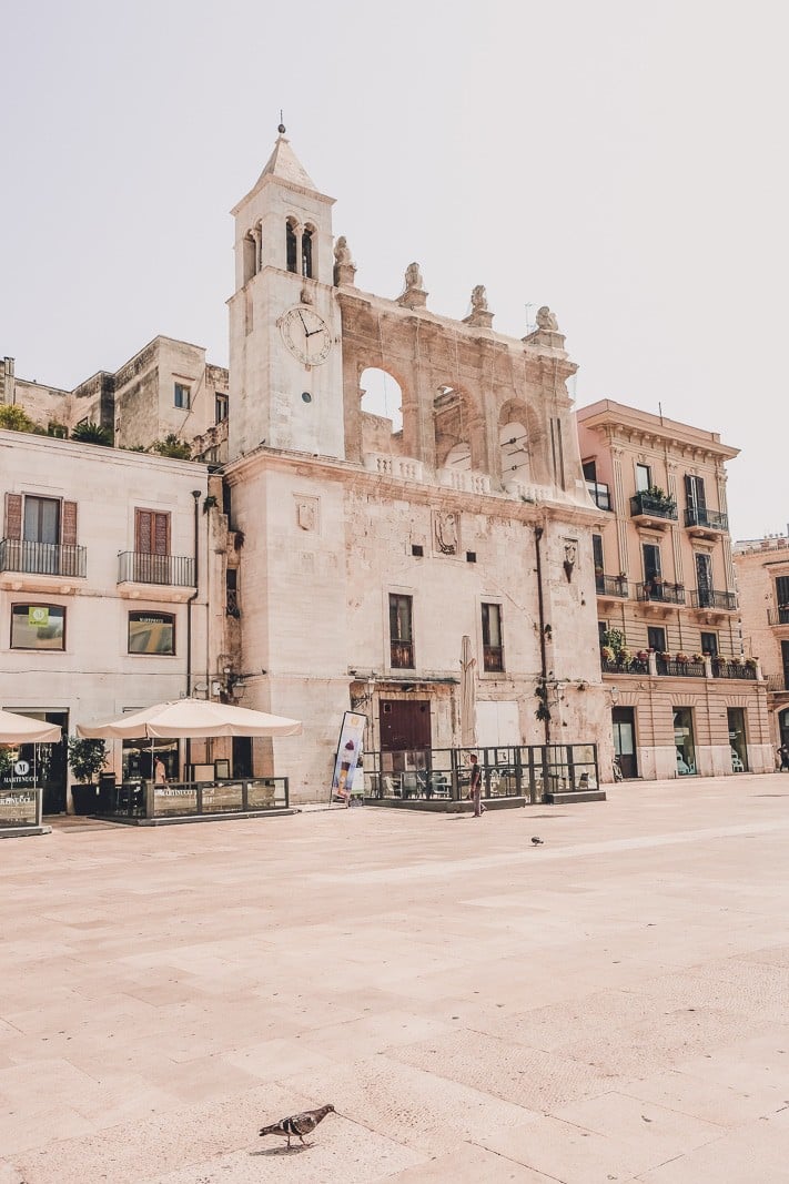 A historic building with a clock tower in Bari, Italy. The building has a worn, white stone facade and a partially ruined upper section with arches. Surrounding the building are modern apartments and outdoor seating areas with umbrellas.