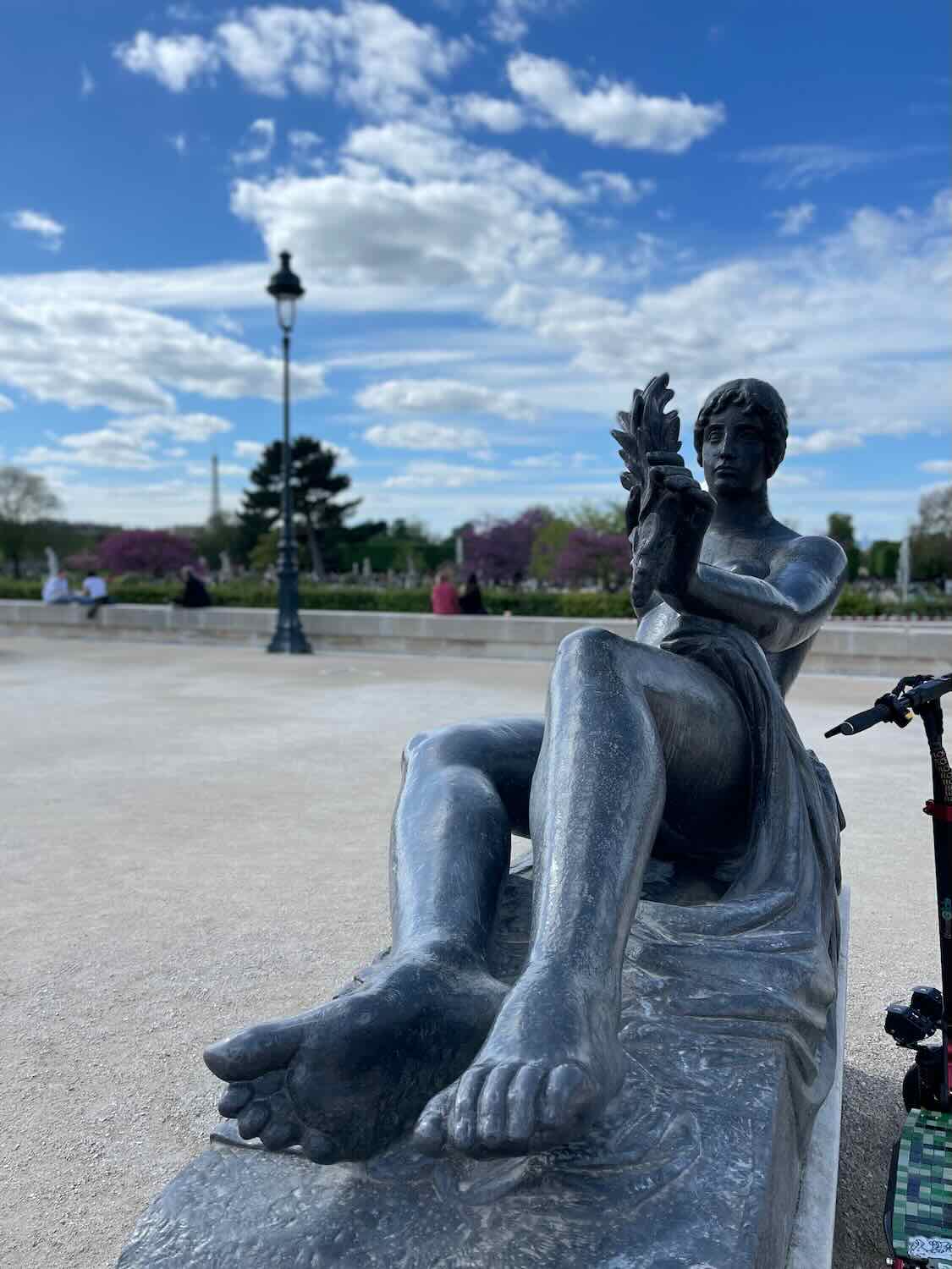 A close-up of a bronze statue of a reclining figure, holding a cluster of leaves, set against a backdrop of Parisian garden scenery with blooming trees and a clear sky.
