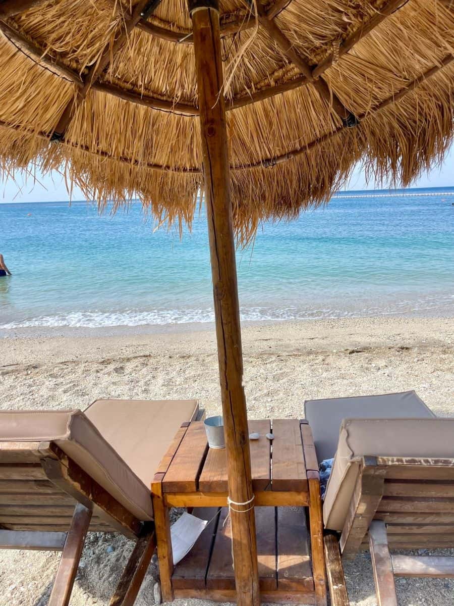 A serene beach scene featuring two lounge chairs under a large thatched umbrella. The chairs face a calm, turquoise sea, with gentle waves lapping the sandy shore. A small wooden table between the chairs holds a few items, and the overall atmosphere is peaceful and relaxing, perfect for a day of unwinding by the water.