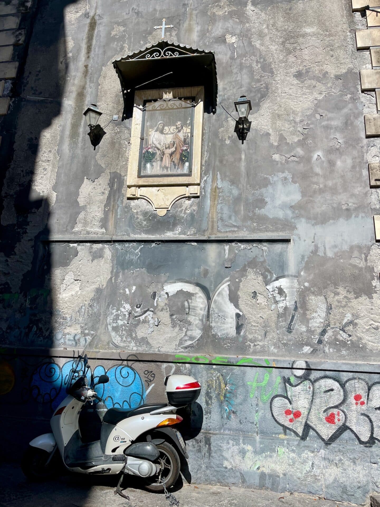 A scooter parked below a religious shrine mounted on a wall with graffiti in Catania.