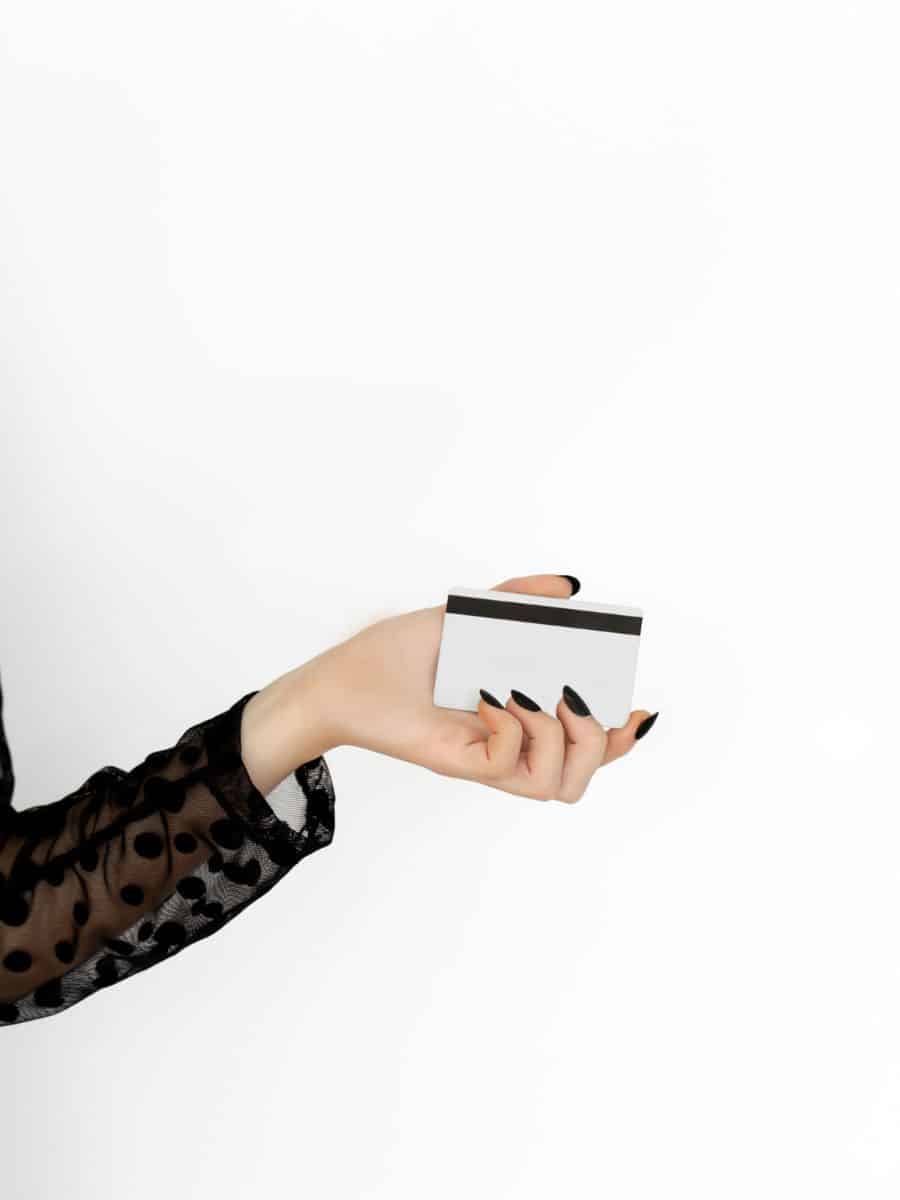 A close-up of a woman's hand with black nail polish holding a blank credit card, wearing a black sheer polka-dot sleeve against a white background.