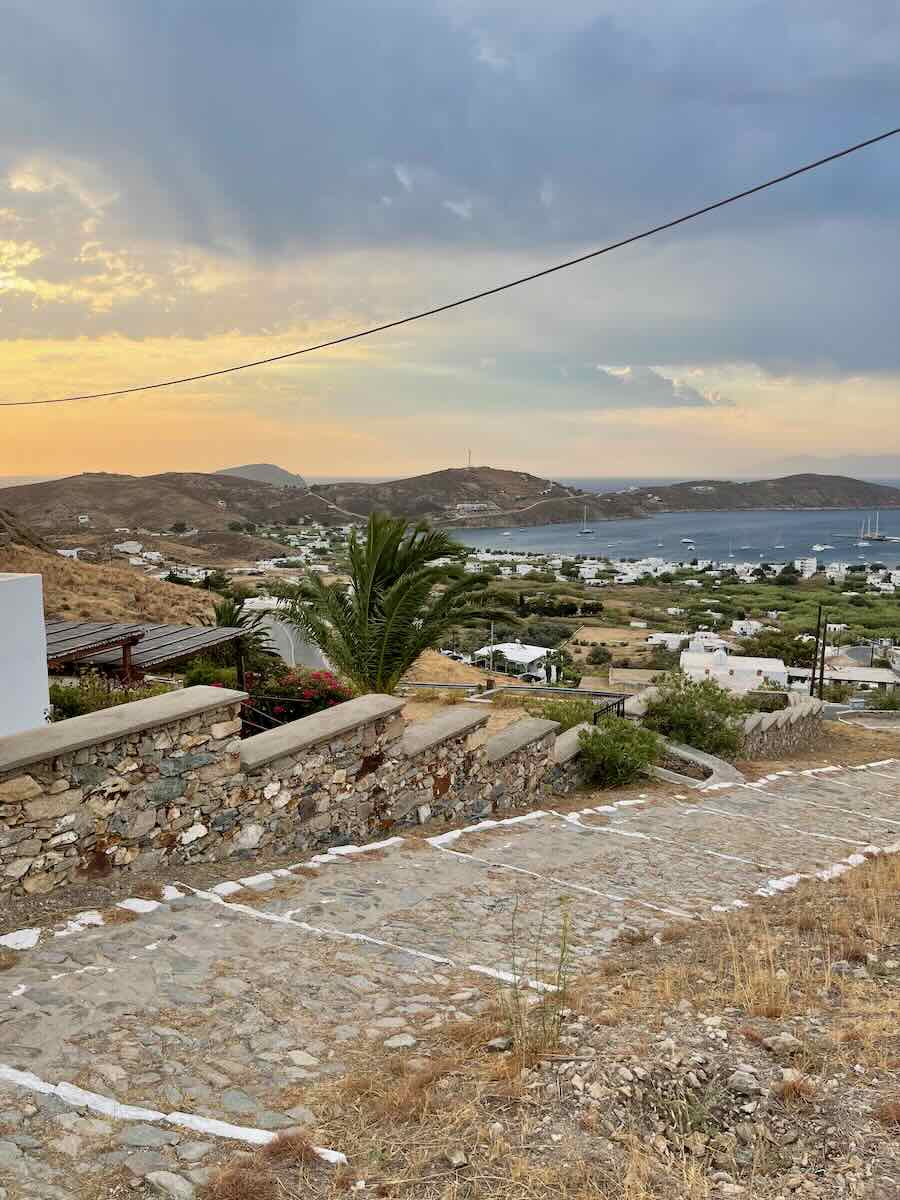 The image shows a stone-paved path leading down a hill toward a coastal village with white buildings. The view overlooks a serene bay with anchored boats and hills in the background. The sky is painted with soft sunset colors, creating a peaceful and scenic atmosphere. A stone wall lines part of the path, and palm trees and other greenery add to the Mediterranean charm of the landscape.