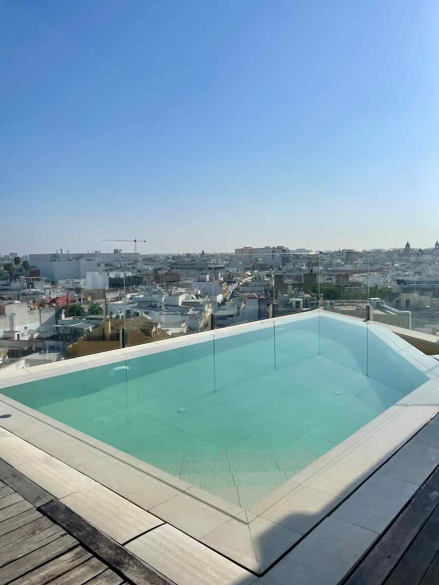 A rooftop swimming pool in Seville, Spain, with clear, calm water and a glass barrier overlooking the city. The view includes the rooftops and skyline of Seville under a cloudless, bright blue sky.