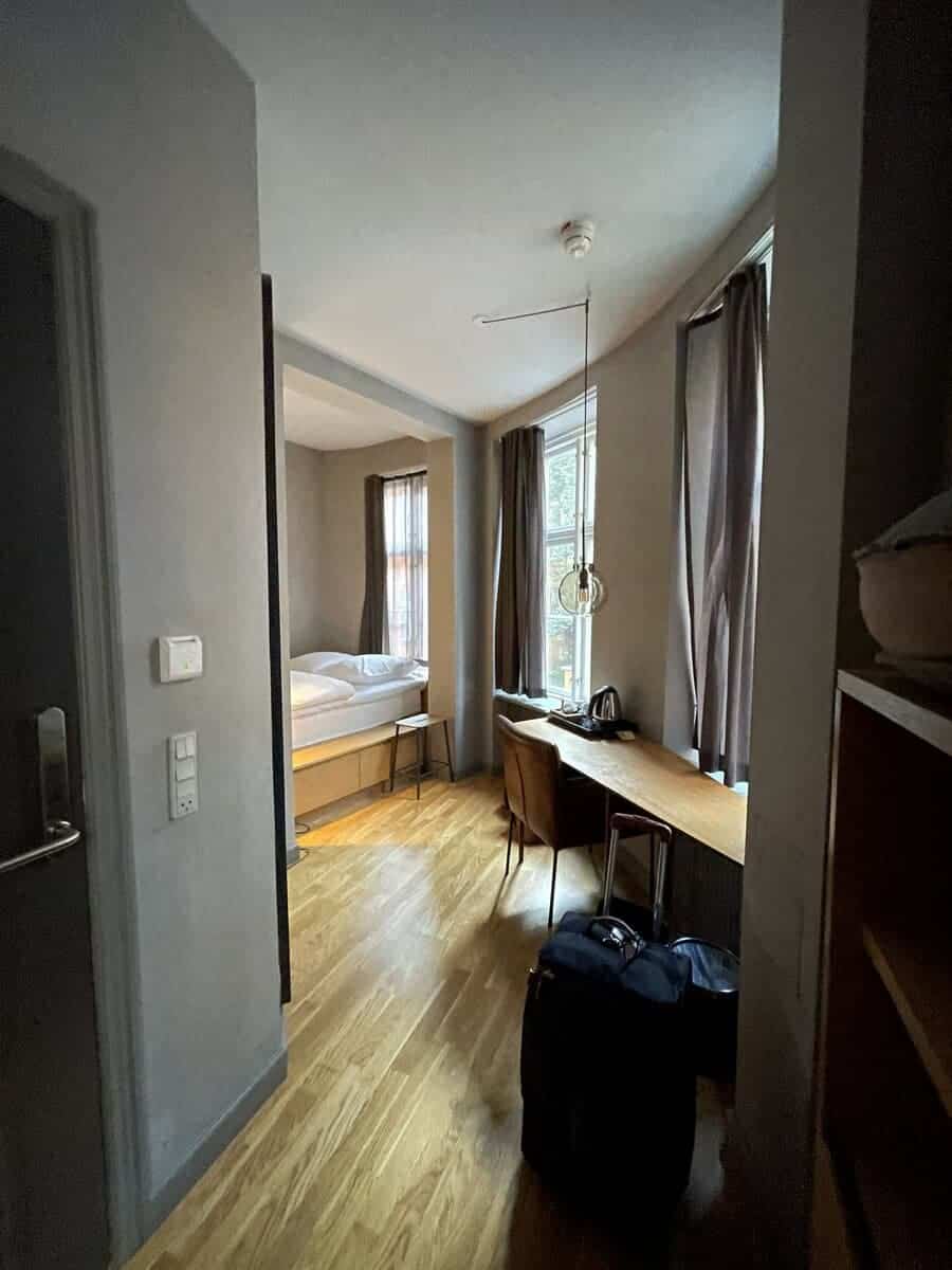 A view into a minimalist room at Hotel SP 34 in Copenhagen, featuring a long wooden desk with a chair, a bed with white linens, and subtle gray curtains over large windows, reflecting a modern and simple decor.