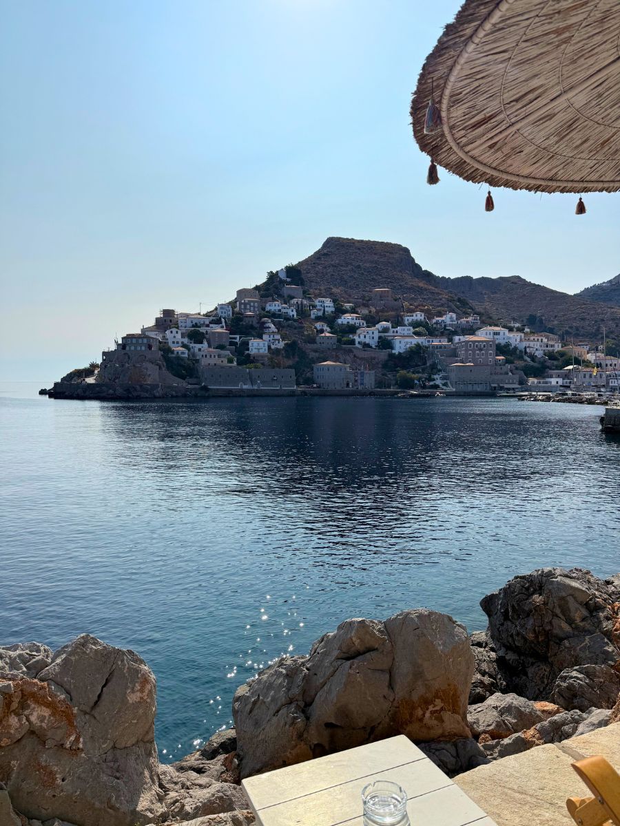 A serene view from a rocky seaside café in Hydra, Greece, showing calm blue waters, a coastal hillside village with white buildings, and part of a straw umbrella above.