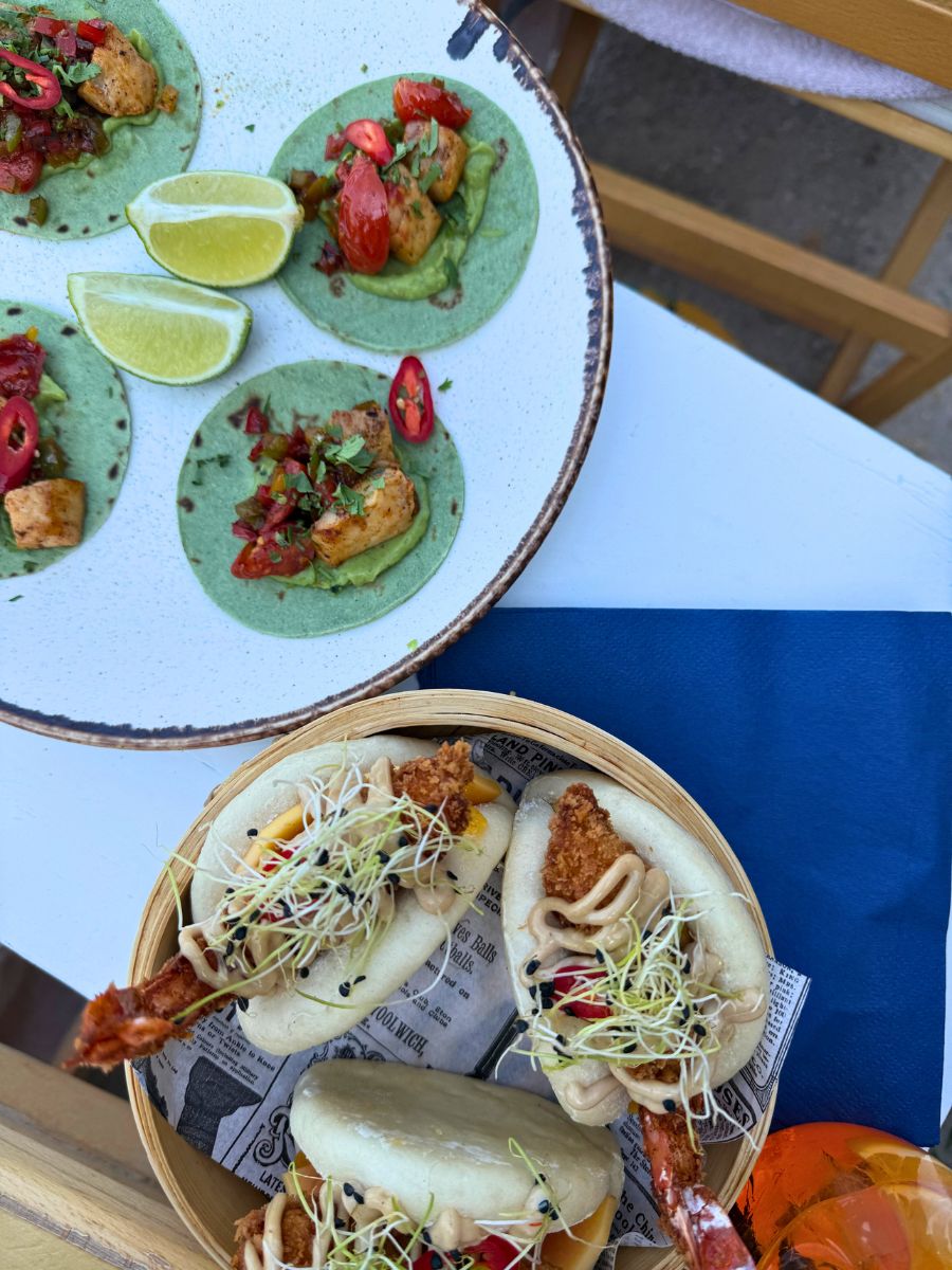 A close-up of a vibrant meal in Hydra, Greece, featuring green mini tacos with grilled chicken, guacamole, cherry tomatoes, and lime wedges, alongside a bamboo steamer basket of crispy shrimp bao buns topped with sprouts and sesame seeds.
