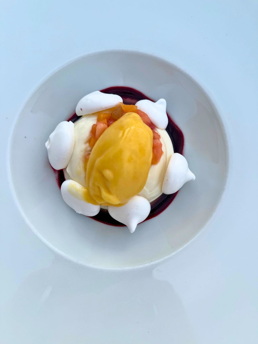 A beautifully plated dessert in Hydra, Greece, featuring a scoop of bright mango sorbet atop a creamy base, surrounded by meringue kisses, diced fruit, and a dark berry sauce on a white dish.