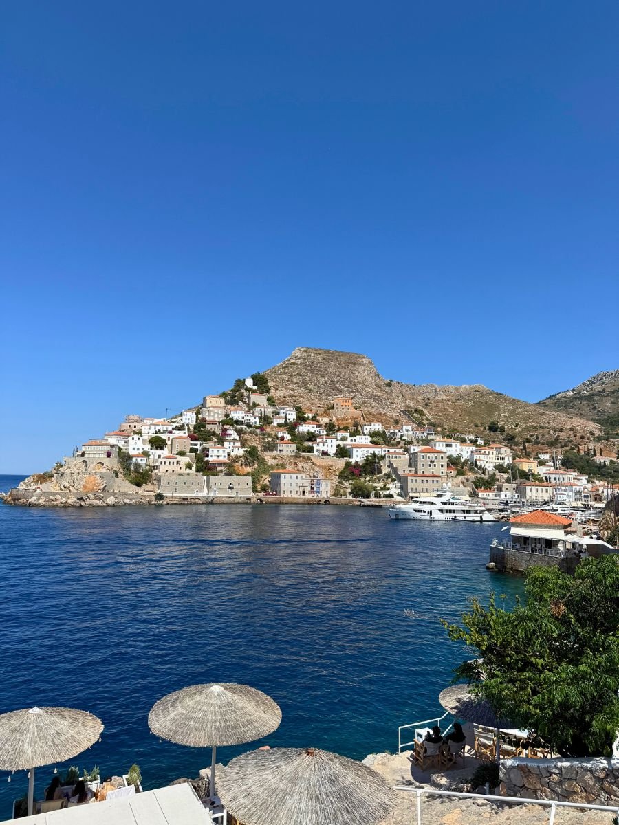 Scenic view of Hydra, Greece, with traditional whitewashed houses scattered on a hillside, overlooking the deep blue Aegean Sea, with straw beach umbrellas and seaside seating in the foreground under a clear blue sky.