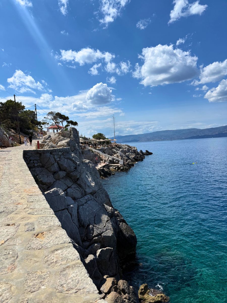 Rocky coastal path on the island of Hydra, Greece, with a stone walkway overlooking the sparkling blue Aegean Sea under a bright sky filled with fluffy clouds.