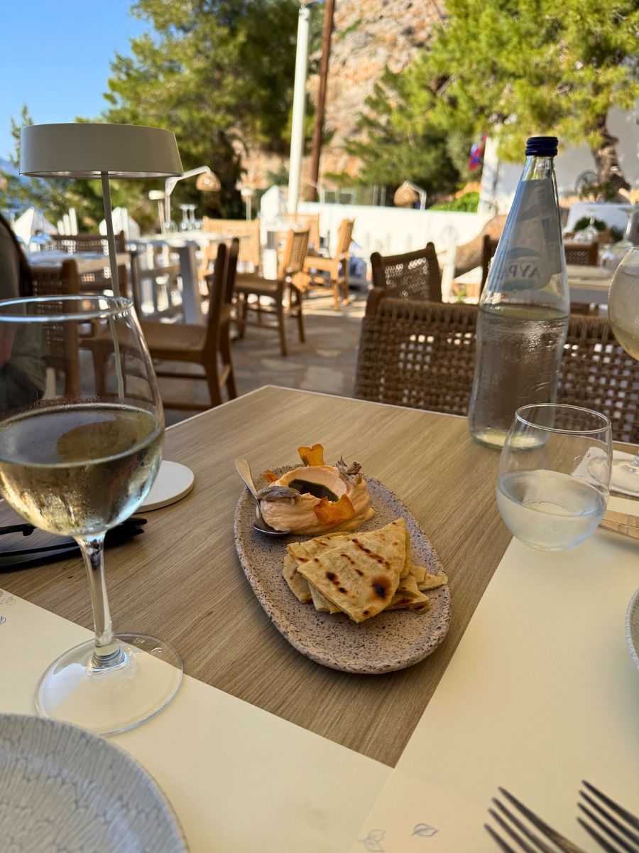 A table set for outdoor dining at a restaurant in Hydra, Greece, featuring a plate of grilled pita bread with dip, a glass of white wine, and a bottle of still water. The background shows a sunlit patio with wooden chairs, white tables, and surrounding trees, creating a relaxed Mediterranean atmosphere.