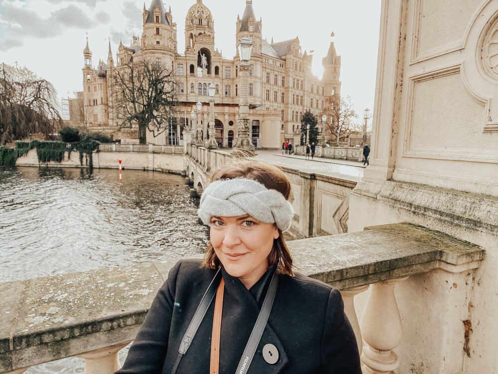 Woman with a headband smiling in front of the historic Schwerin Castle in Germany, illustrating the charm of solo winter travel.