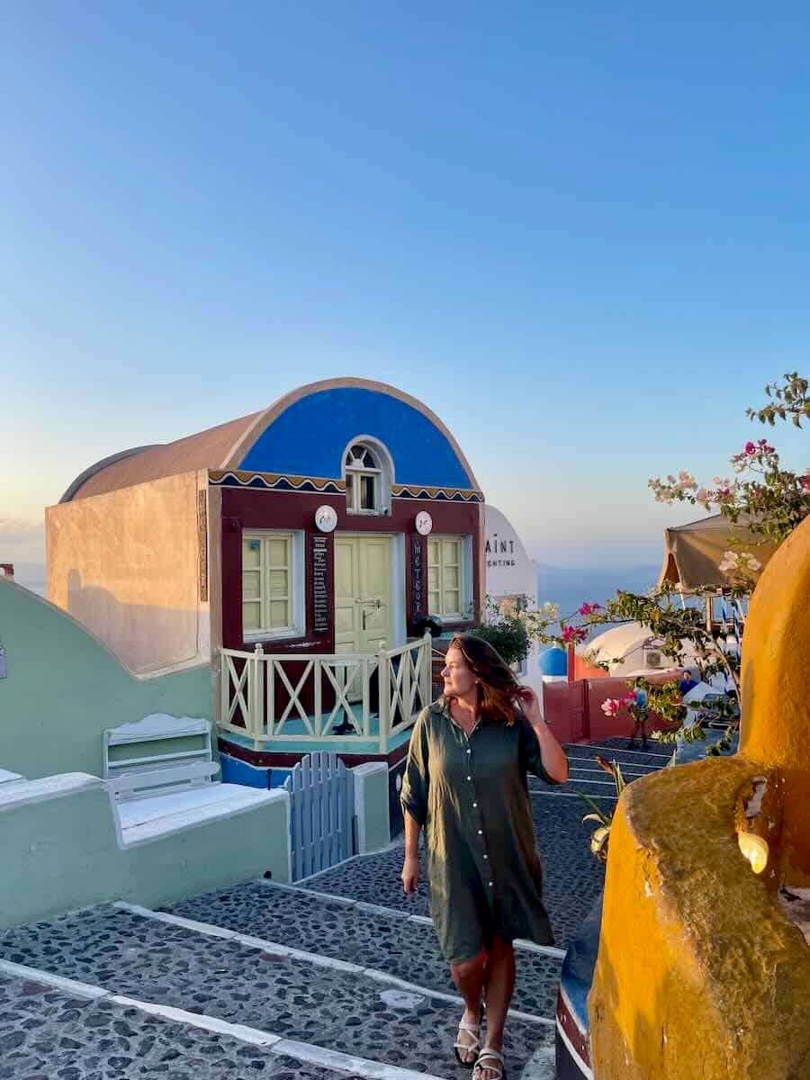Woman walking down a cobblestone street in Santorini, wearing a green dress, with traditional colorful houses and a clear blue sky in the background at sunset.