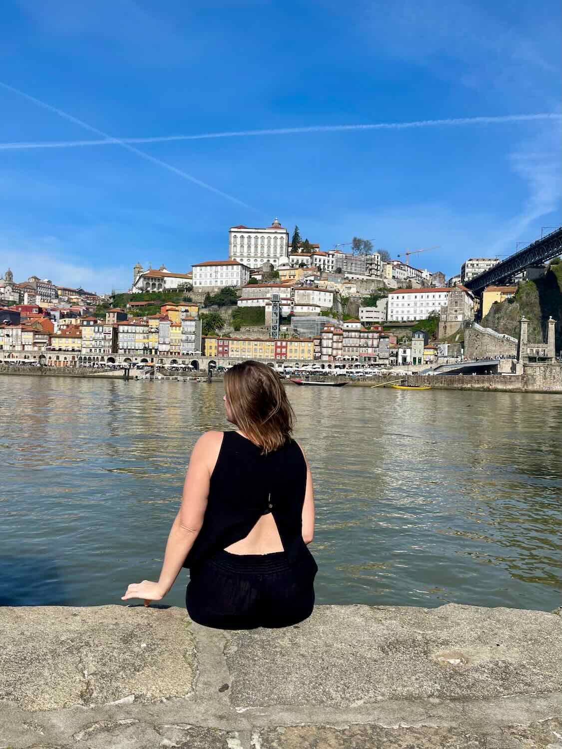 A woman sits on the edge of a stone wall by the Douro River in Porto, gazing across at the colorful buildings of the city on the opposite bank. The hillside is dotted with vibrant houses, and the iconic Dom Luís I Bridge is visible to the right. The clear blue sky and reflections on the water create a peaceful, picturesque scene of Porto’s riverside charm.