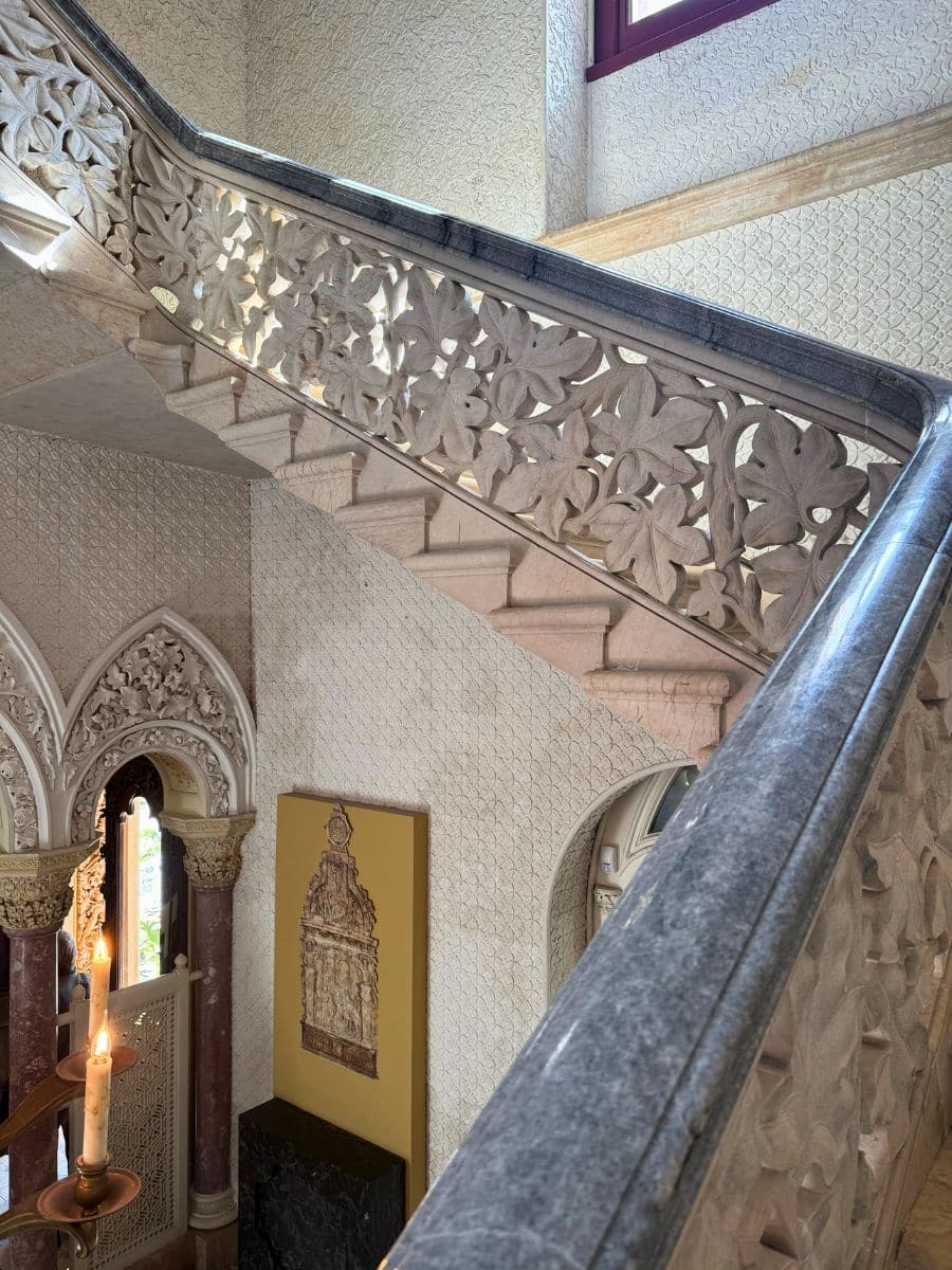 Intricately carved stone staircase inside a palace in Sintra, featuring floral patterns on the banister and arched Gothic-style doorways with decorative molding and candle sconces.