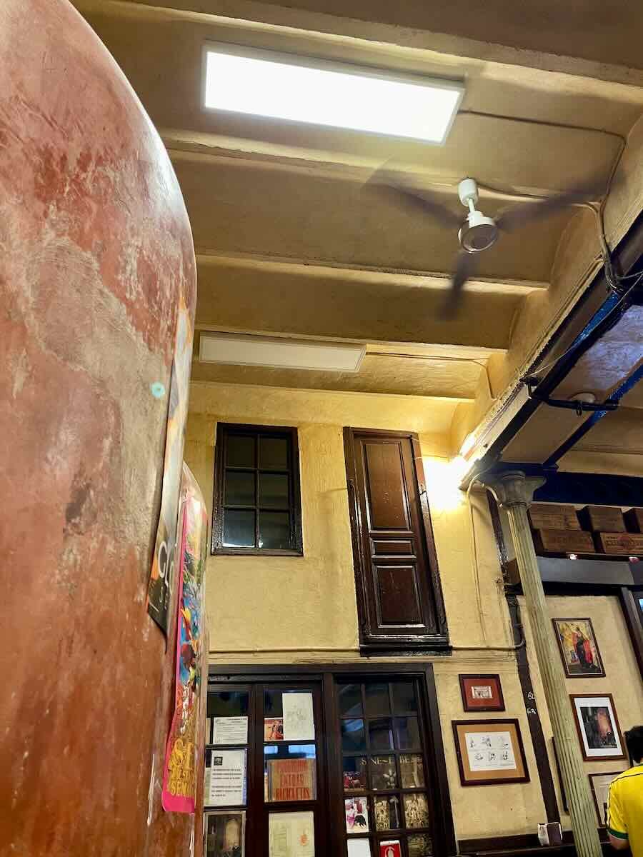 An interior shot of Casa Morales, a traditional bar in Seville, Spain. The image captures part of a large clay wine jar, a ceiling fan in motion, and walls adorned with framed photos and posters. The bar’s rustic decor, with exposed beams and vintage touches, is visible.