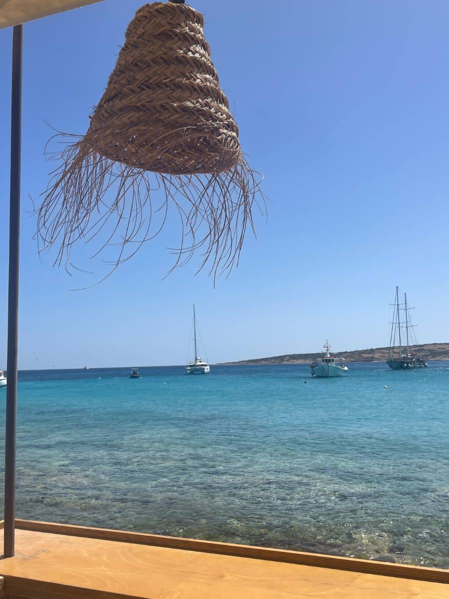 A rustic woven lampshade sways in the breeze above a wooden beachside table, overlooking sailboats anchored in the calm blue sea.