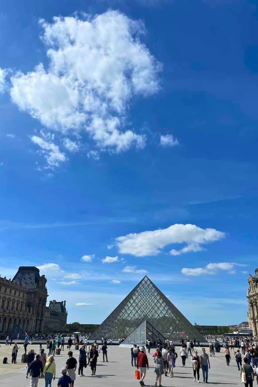 clear day scene at the Louvre Museum's courtyard, showcasing the large glass pyramid under a blue sky scattered with clouds. 