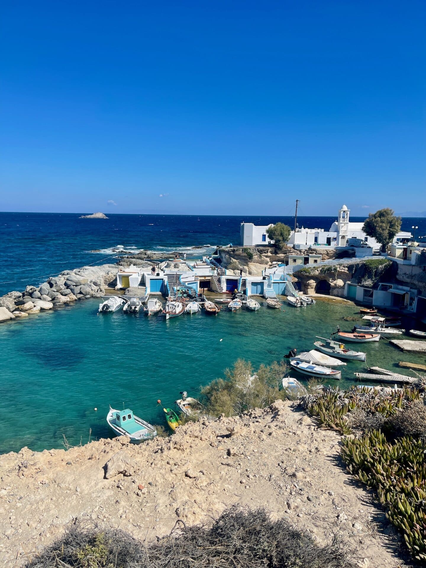 A picturesque view of a small coastal village with white buildings and a serene turquoise bay. Several boats are docked in the calm water, and the clear blue sky meets the horizon where the sea stretches out. The landscape has rocky terrain in the foreground and the peaceful, charming village sits by the sea.