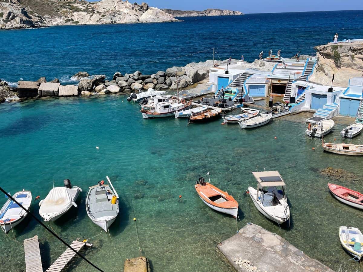 The image shows a small, clear turquoise harbor with several boats anchored along the shore. Blue and white boathouses line the edge of the water, giving the scene a charming, coastal vibe. The deep blue sea stretches beyond the harbor, with rocky cliffs in the background.