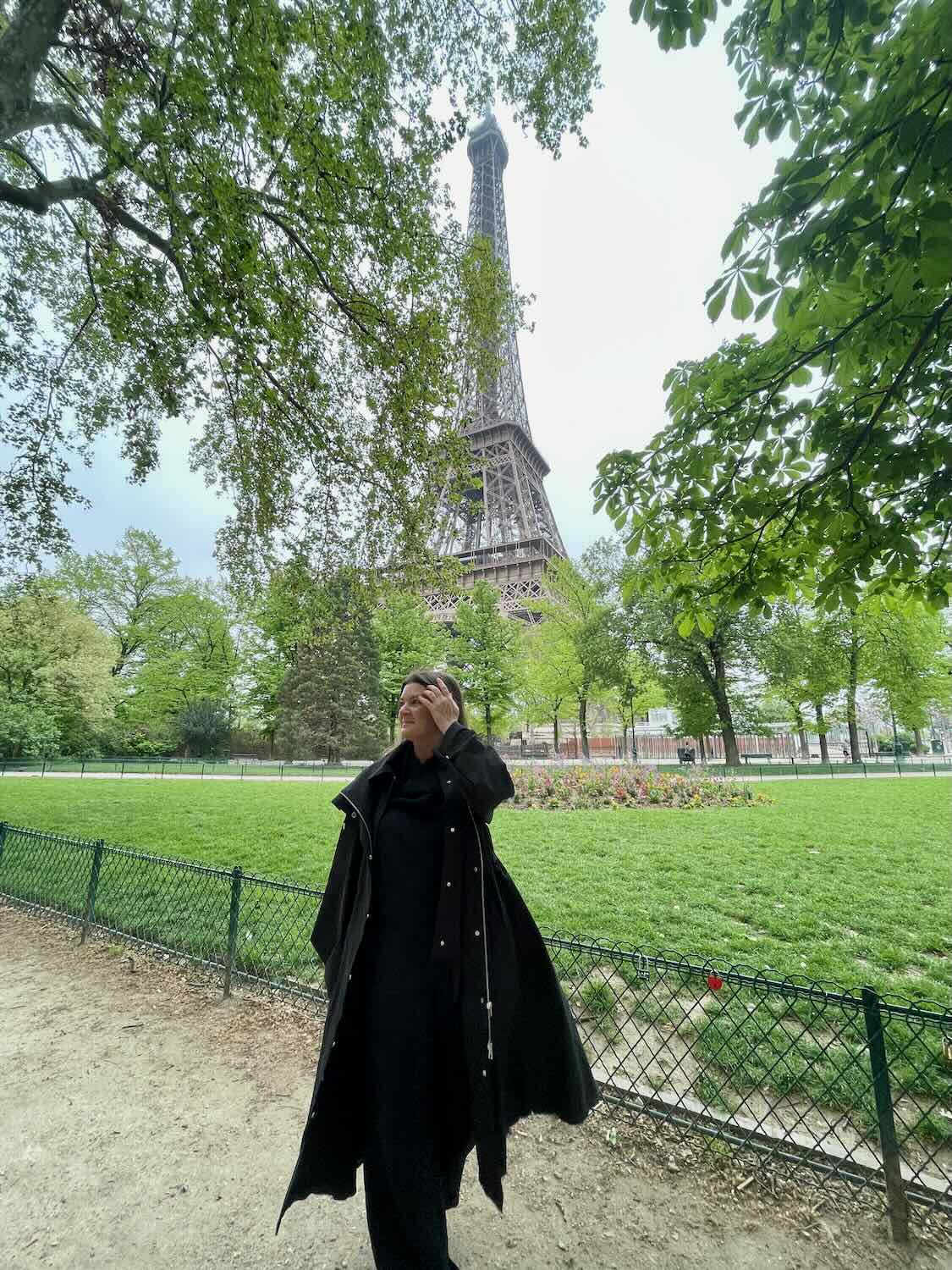 A solo female traveler stands in a park near the Eiffel Tower, dressed in an elegant all-black outfit with a long coat. She looks away thoughtfully, touching her hair, as the iconic tower rises majestically in the background, framed by lush green spring foliage.