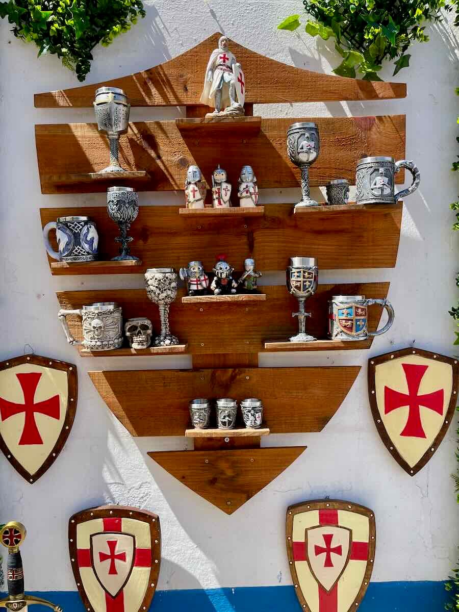 A wooden wall display in Óbidos, adorned with medieval-style goblets, figurines of knights, and shields bearing the red cross of the Knights Templar. This display gives a glimpse into the medieval culture of the town.