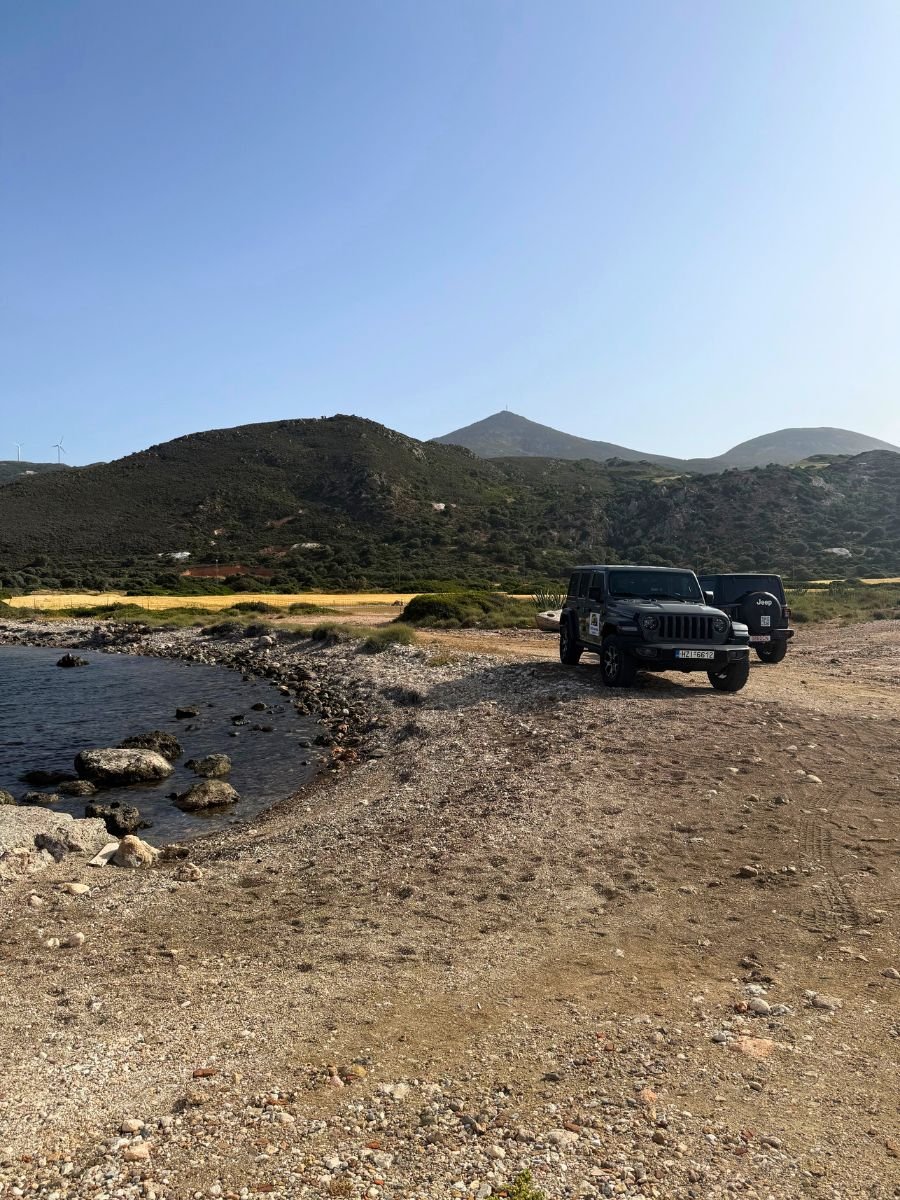 A rugged coastal landscape in Milos, Greece, showing a rocky shoreline with dark waters meeting a pebbly, dirt path. Two black Jeep vehicles are parked on the right, facing the mountains that rise in the background under a clear blue sky. Sparse vegetation dots the area, and a line of wind turbines can be seen in the distance on a ridge.
