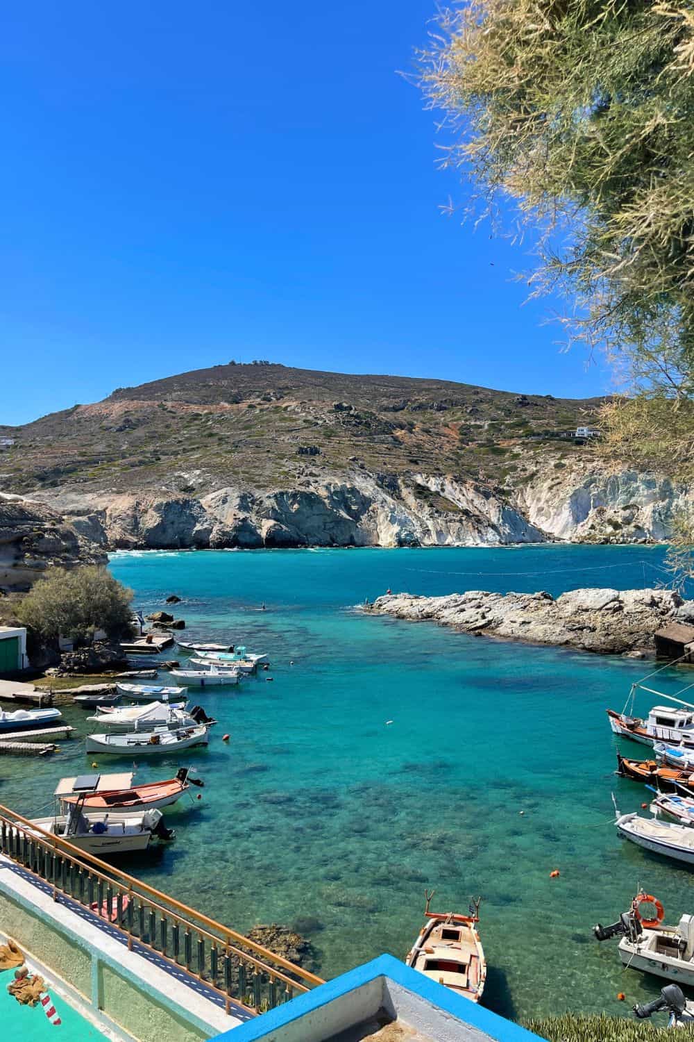 Scenic view of a turquoise bay lined with boats, surrounded by rugged hills, under a clear sky.