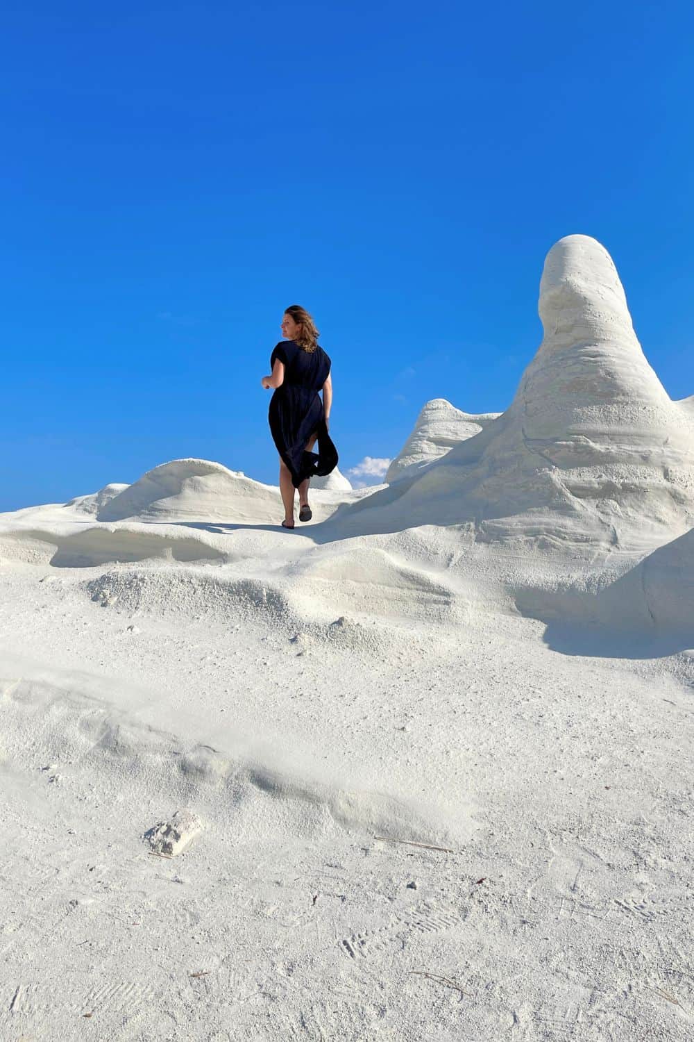 Woman in a black dress walking on white, moon-like rock formations under a clear blue sky.