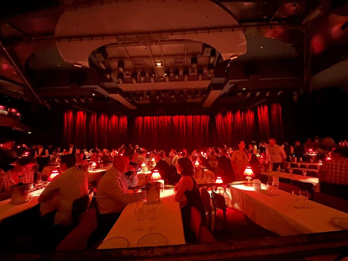 vibrant and atmospheric setting of a Parisian cabaret. The venue is bathed in a warm red light, featuring a large stage with red curtains and tables spread throughout the space where patrons sit enjoying the show. 