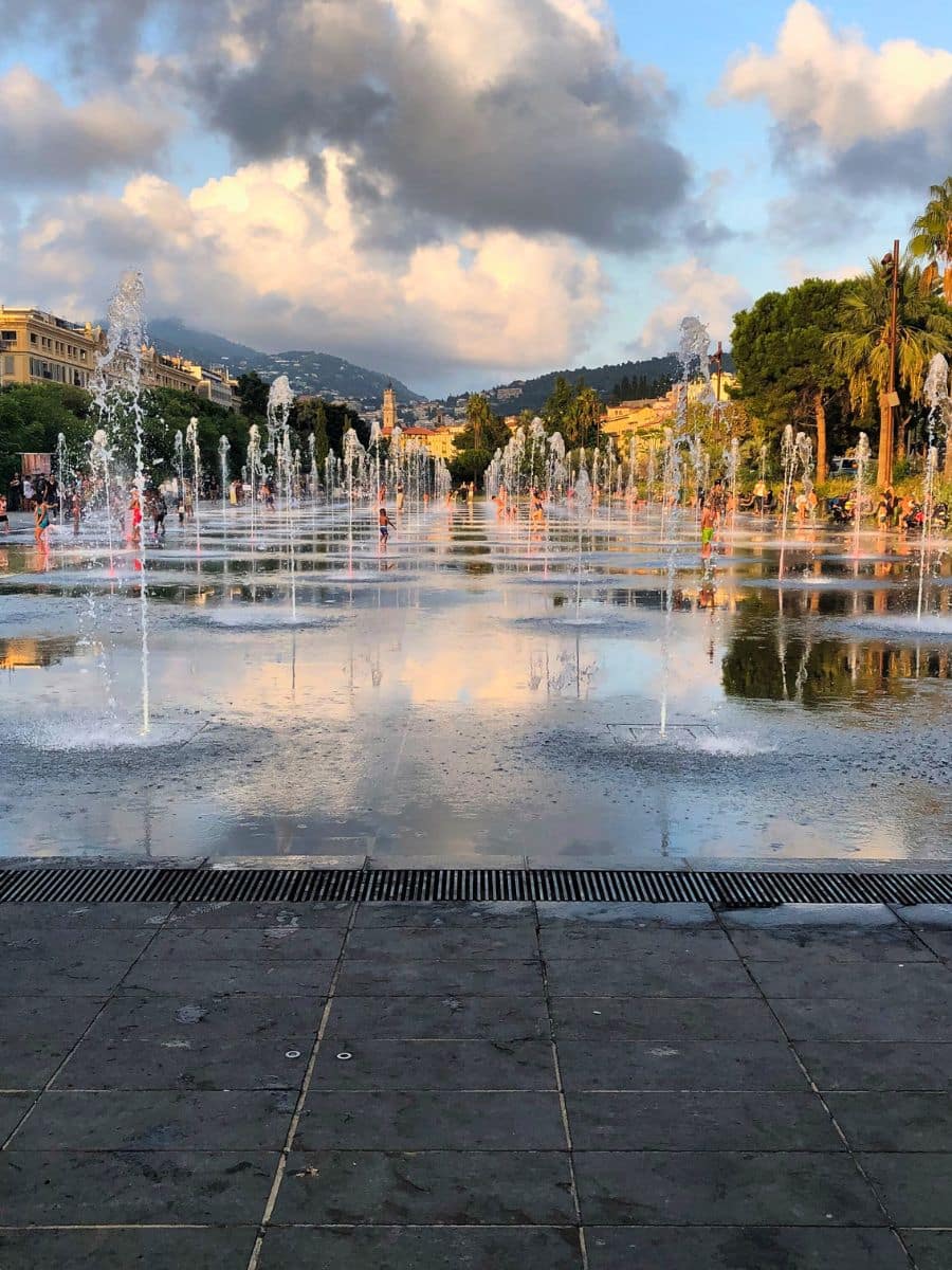 Dynamic scene of children playing in a water mirror fountain, with jets of water rising around them, against a backdrop of palm trees and mountains at sunset.